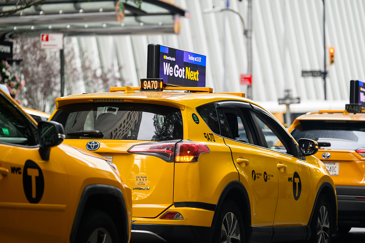 Multiple NYC yellow taxis with digital LED advertising screens on their rooftops, displaying Best Buy ads in a busy Manhattan traffic scene